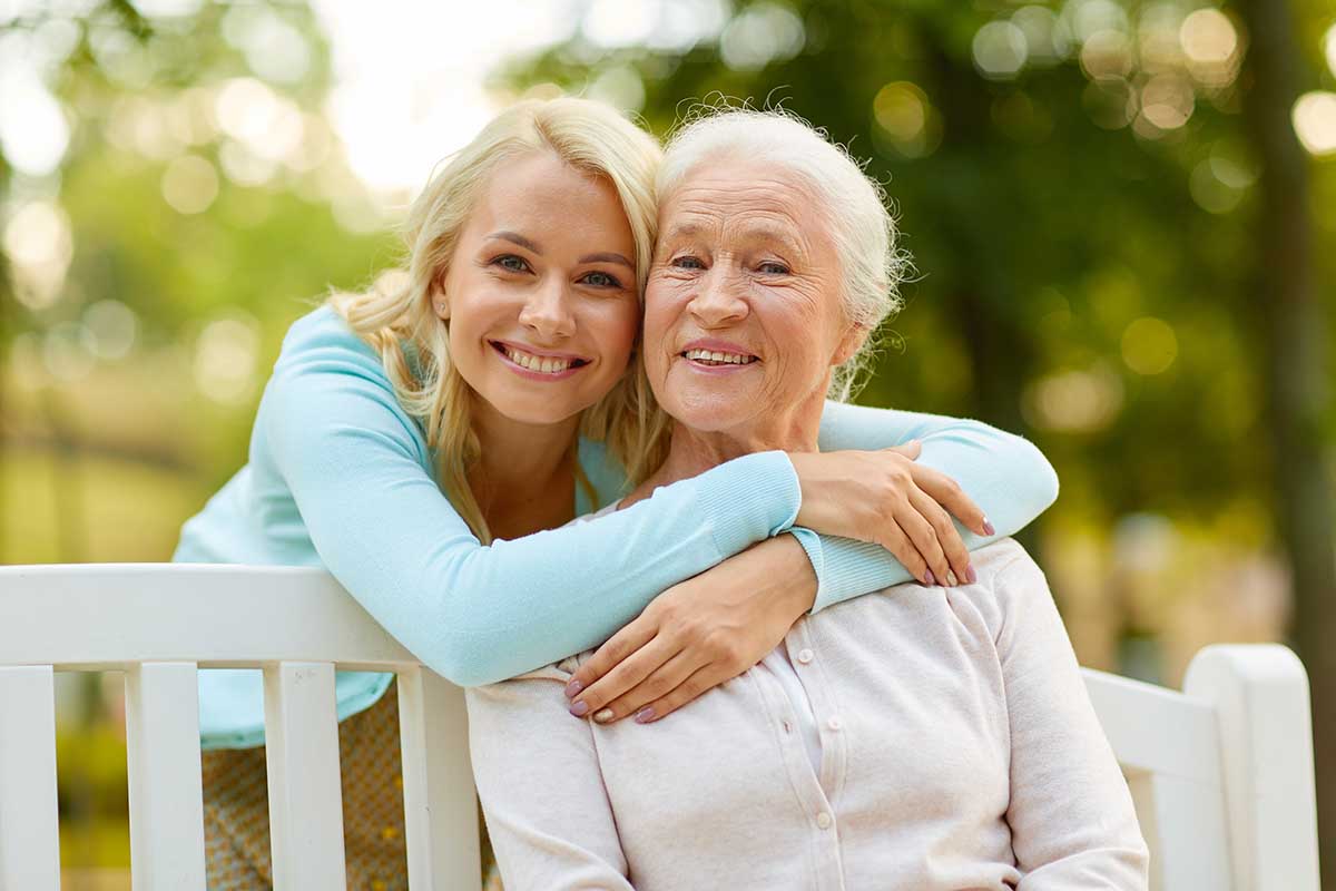 happy young woman hugging her smiling grandmother sitting on a bench in a park