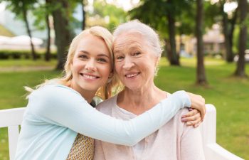 happy young woman hugging her smiling grandmother sitting on a bench in a park