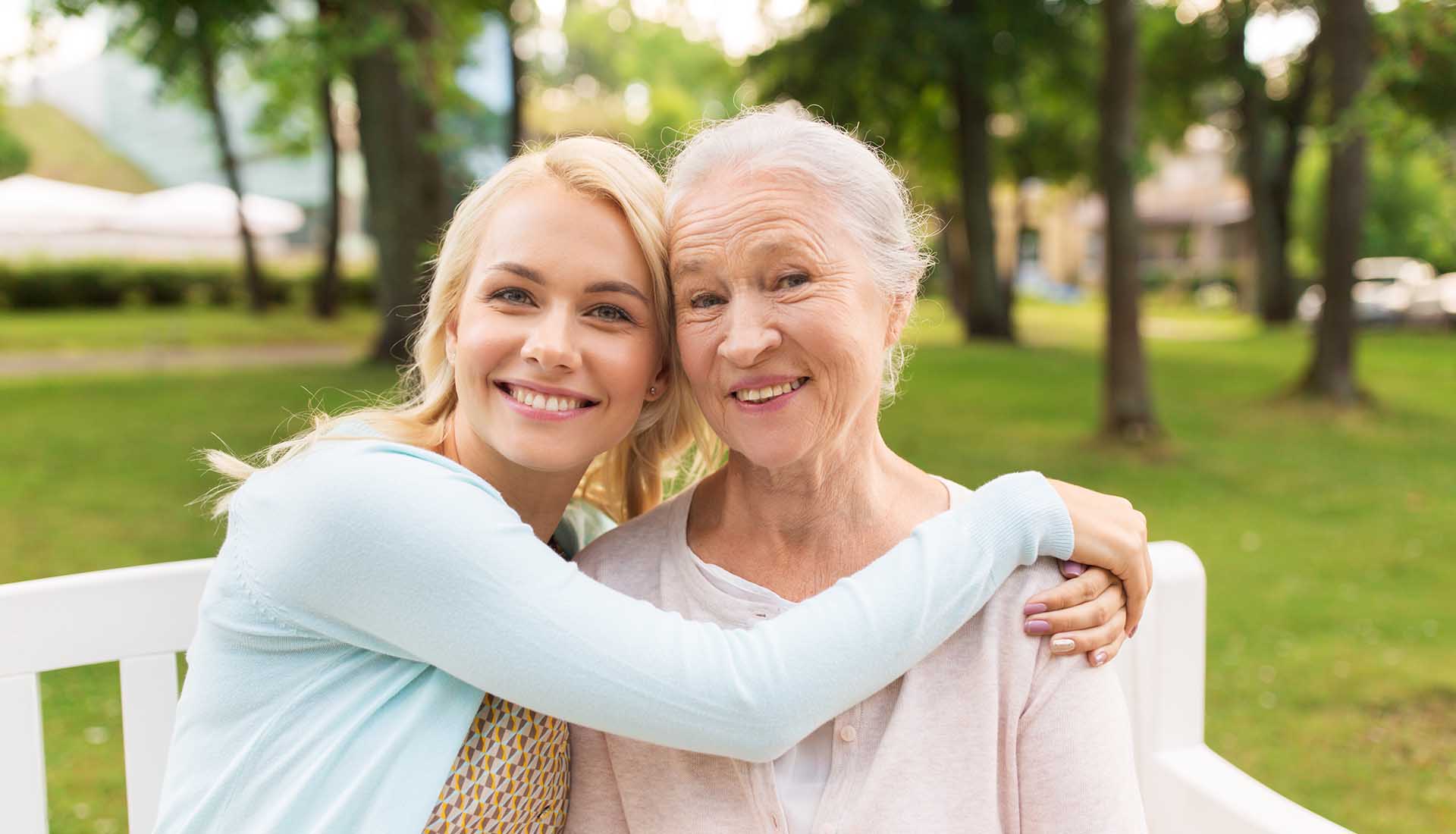 happy young woman hugging her smiling grandmother sitting on a bench in a park