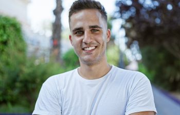 A man wearing a white t-shirt stands outdoors, smiling at the camera, with greenery and trees in the blurred background.